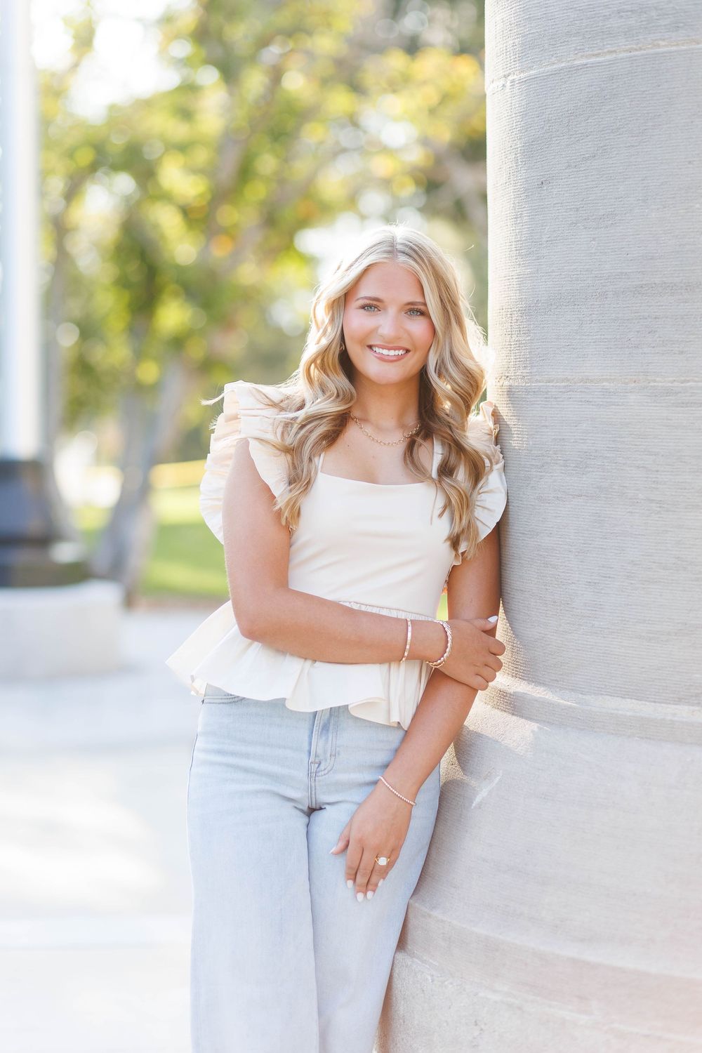 A person in a white sleeveless top and light jeans poses against a white wall in a sunny outdoor setting.