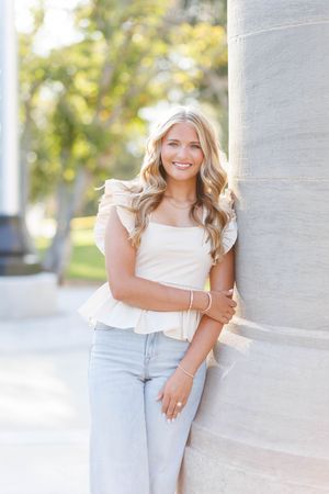 A person in a white sleeveless top and light jeans poses against a white wall in a sunny outdoor setting.
