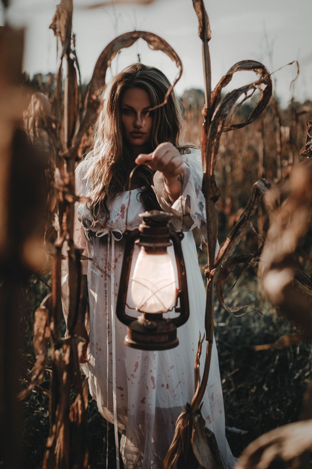 A figure in a white dress holds an antique lantern in a moody forest setting at dusk.