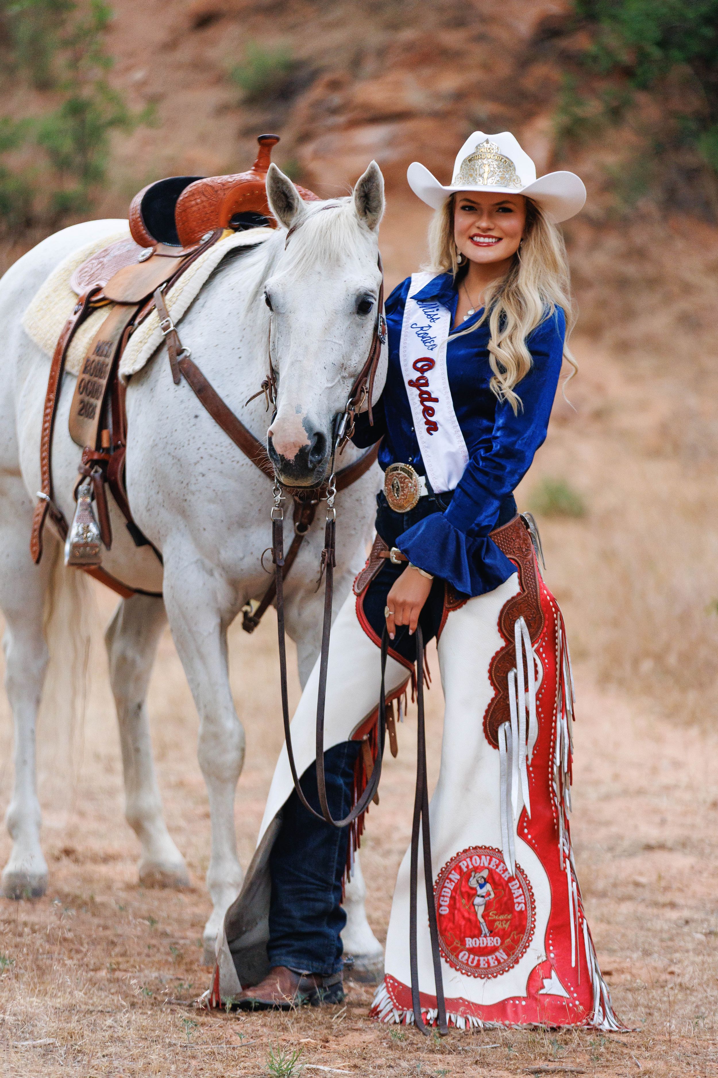 A smiling rodeo queen in blue velvet jacket, white cowboy hat, and decorative chaps poses beside a white horse outdoors.