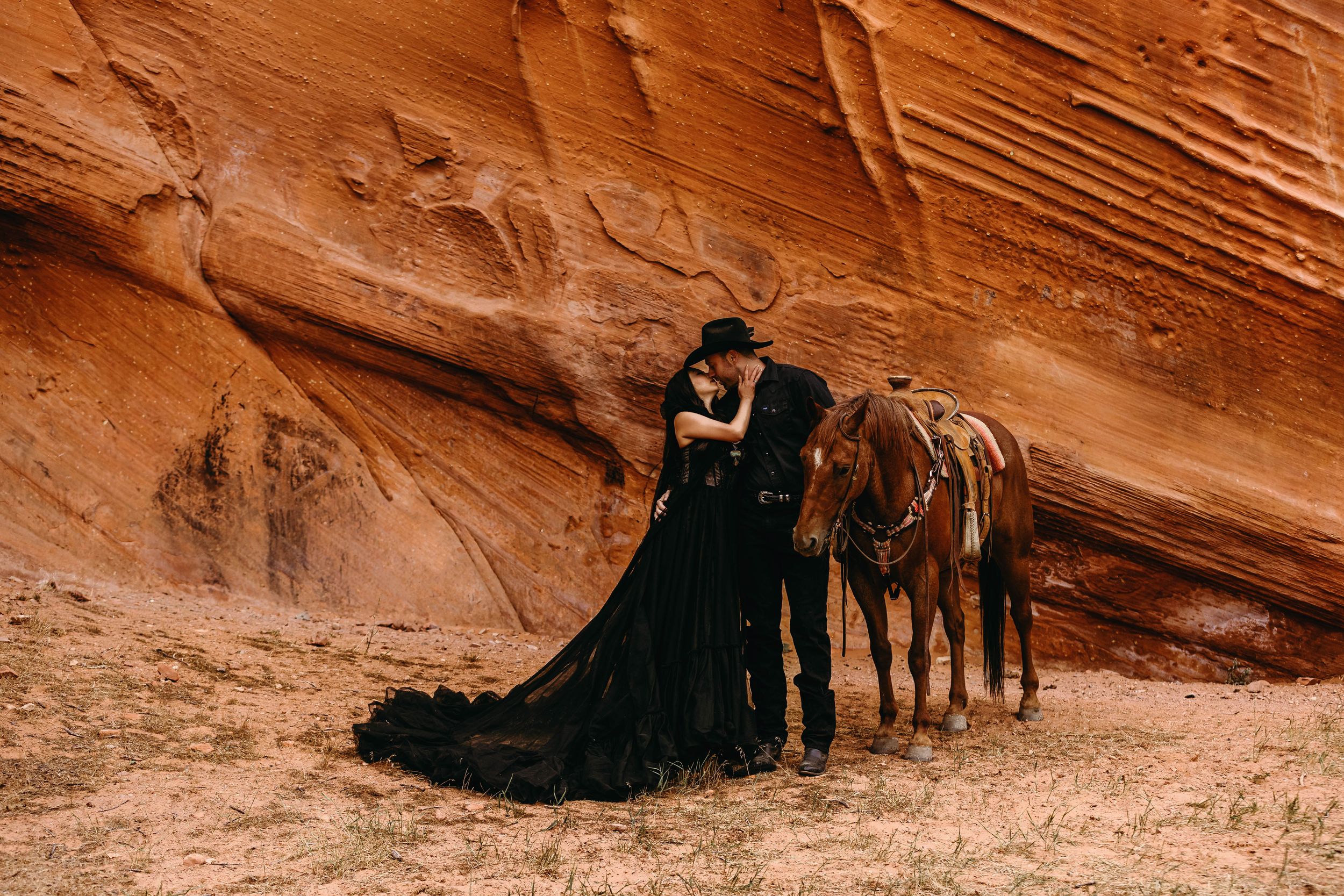 Couple in black attire embracing near a horse in a stunning red rock canyon desert landscape.