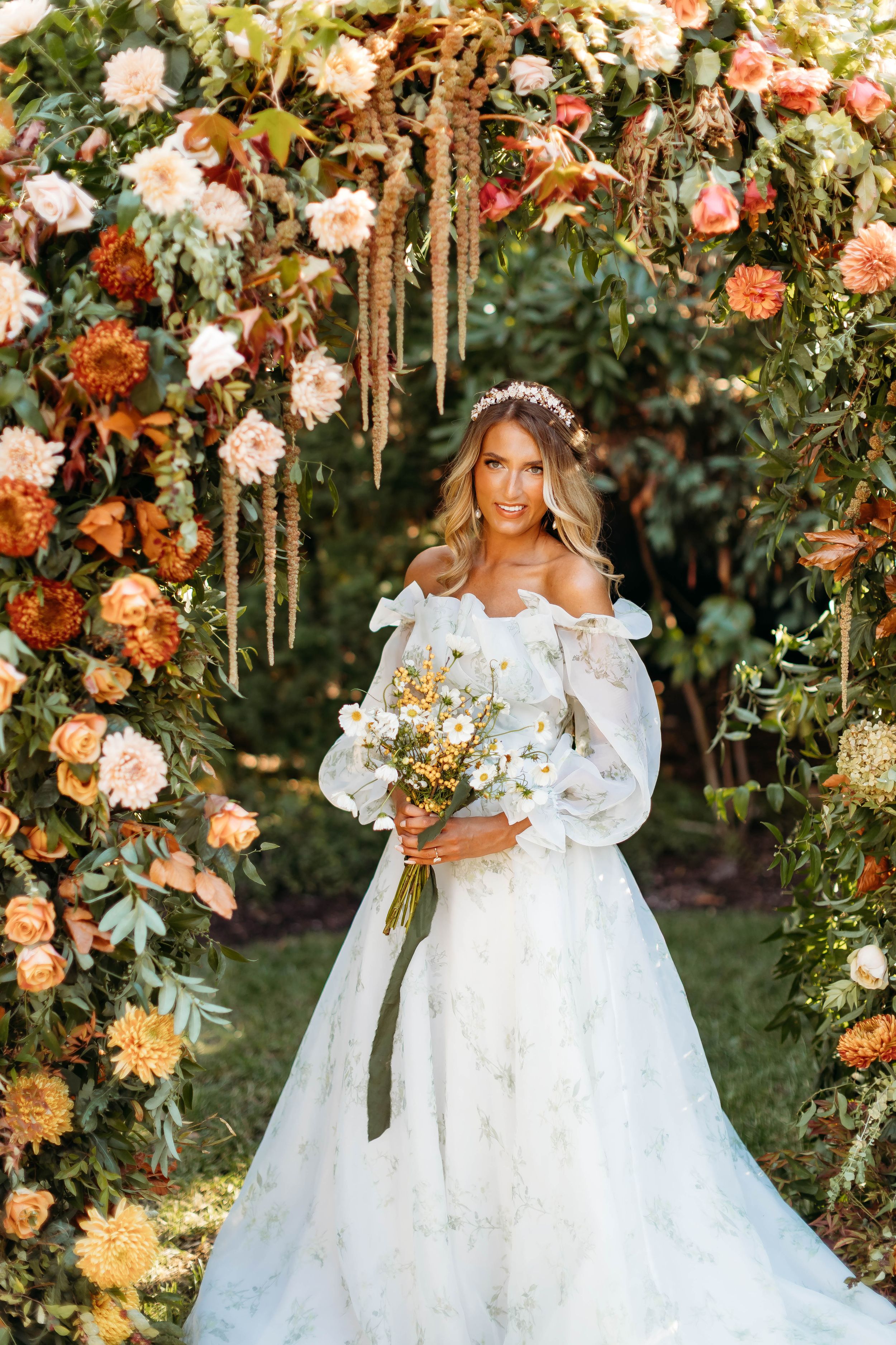 Bride in off-shoulder floral gown holding white bouquet under autumn flower arch with orange and pink blooms.
