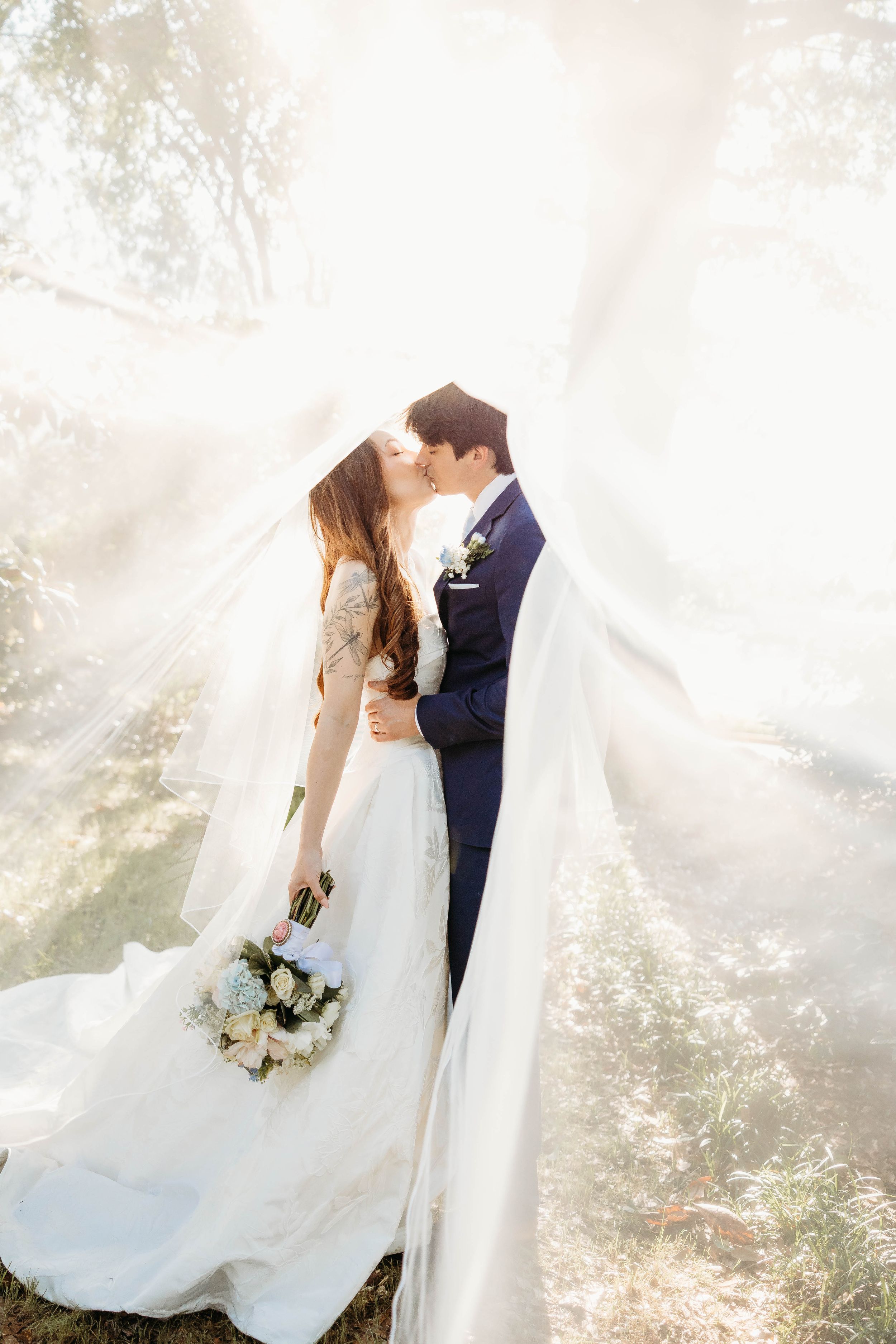 Bride and groom share a romantic kiss outdoors, wrapped in a flowing white veil with soft sunlight streaming through.