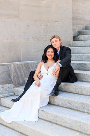 A couple in formal attire sits on stone steps, the woman in a white gown leaning against the man in a dark suit.