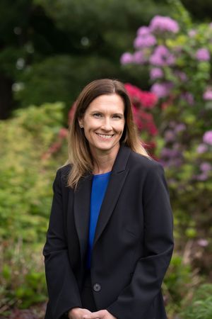 Professional portrait of someone in a black blazer and blue top standing outdoors with pink flowers in background.
