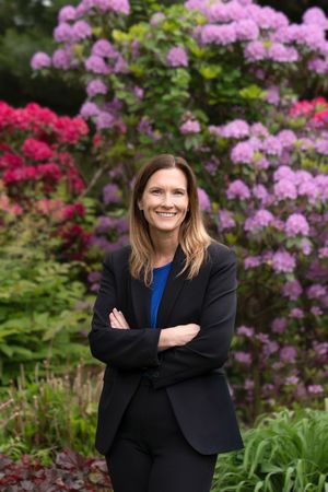 Professional in a black blazer and blue blouse poses with crossed arms in front of vibrant pink rhododendron flowers.