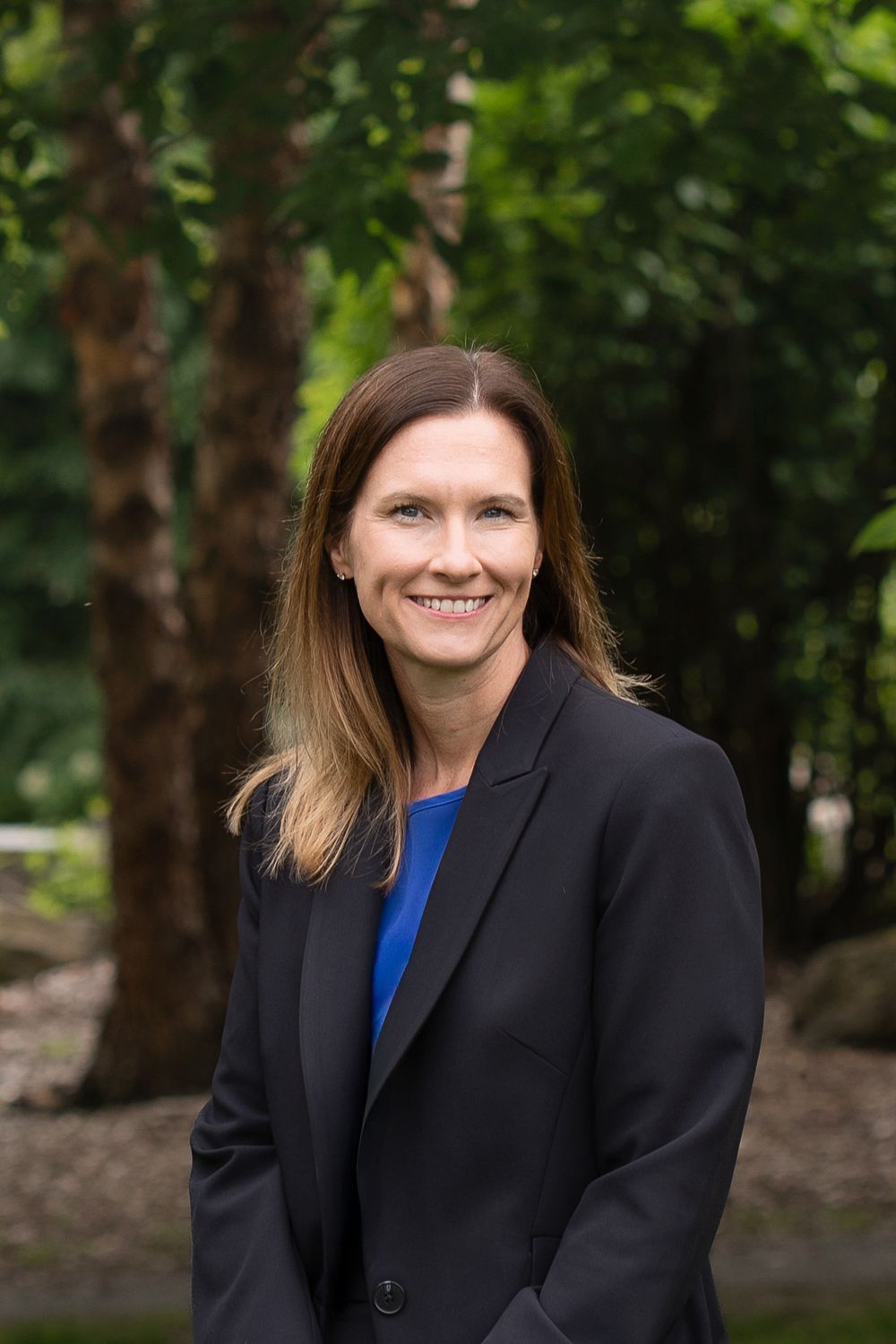 Professional headshot of person in black blazer and blue top standing outdoors with green foliage background.