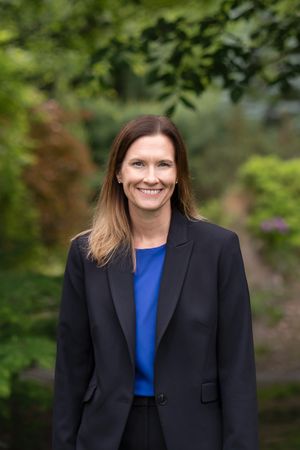 Professional portrait of someone in a black blazer and blue top standing outdoors with greenery in the background.