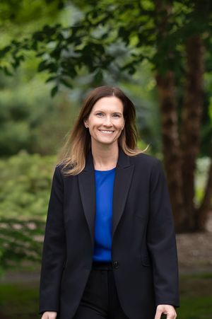 Professional office portrait outdoors showing a person in a navy blazer and vibrant blue top against a blurred natural background.
