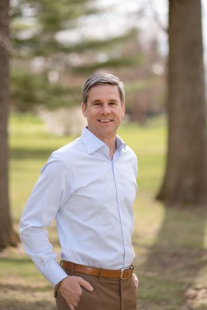 Professional portrait of someone in a light blue dress shirt and khaki pants standing outdoors in a natural setting.