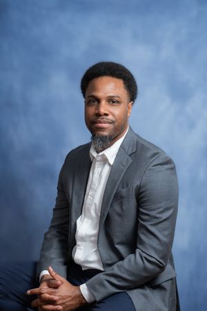 Professional studio portrait against blue backdrop showing confident pose in grey suit.