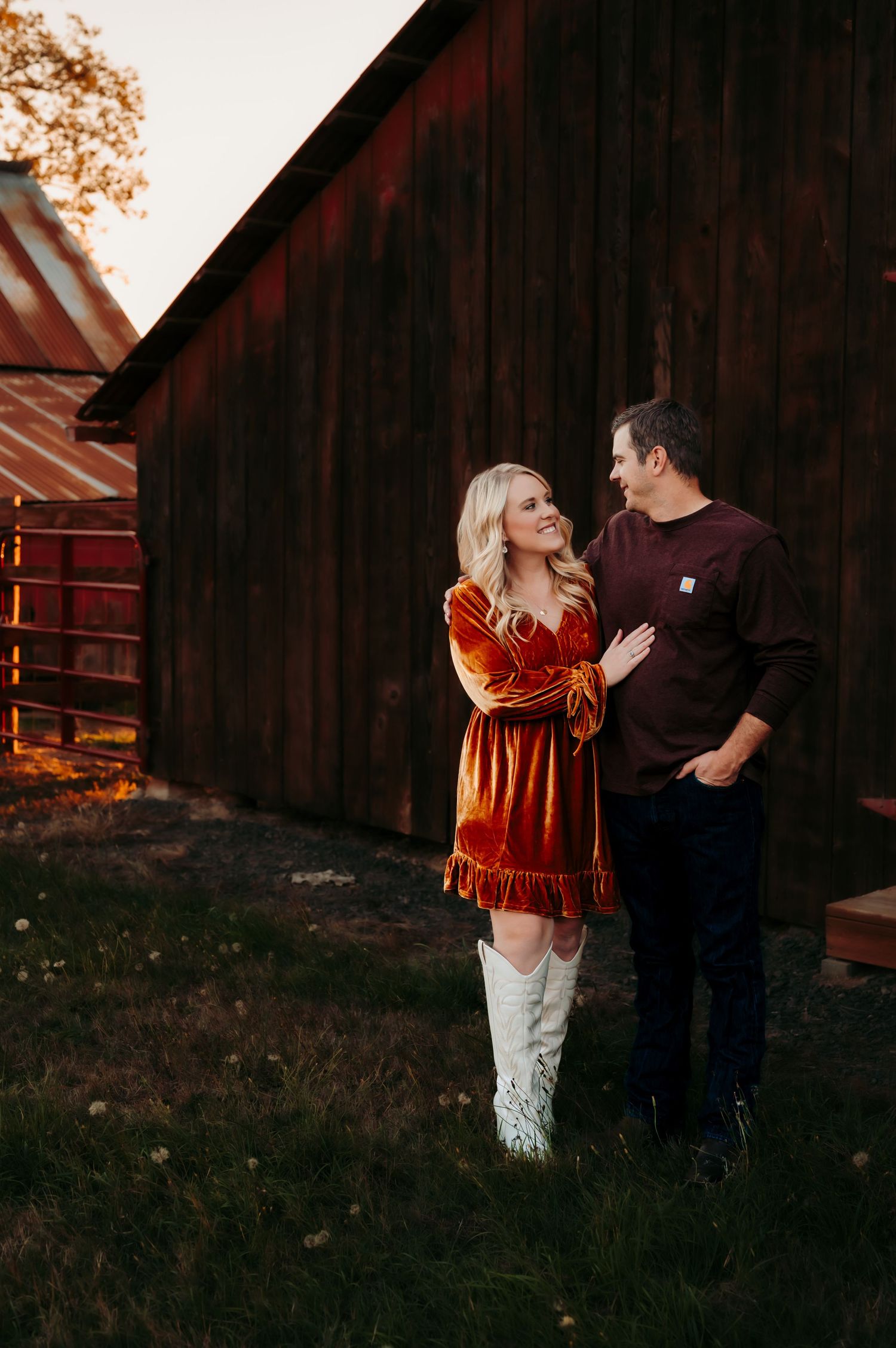 Couple in casual attire poses together against rustic barn backdrop during golden hour.