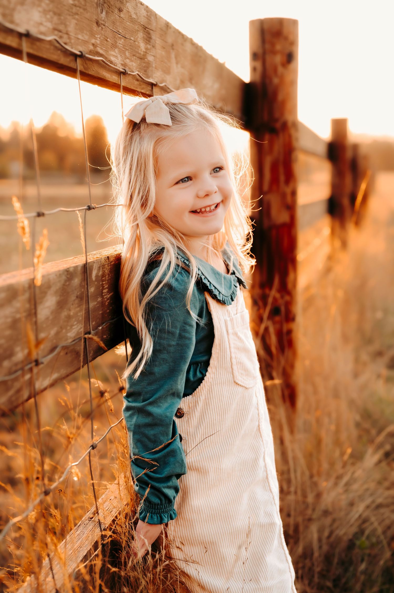 Young child in overalls smiles brightly while leaning against rustic wooden fence at sunset.