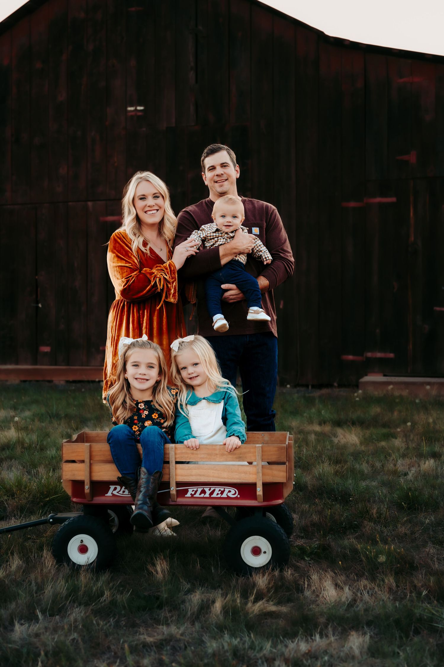 Family poses for portrait by rustic barn with children sitting in red wagon.