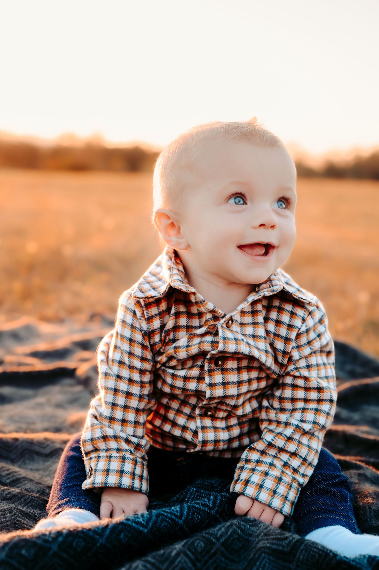 Baby in plaid shirt sits on blanket during golden hour outdoor photo session.