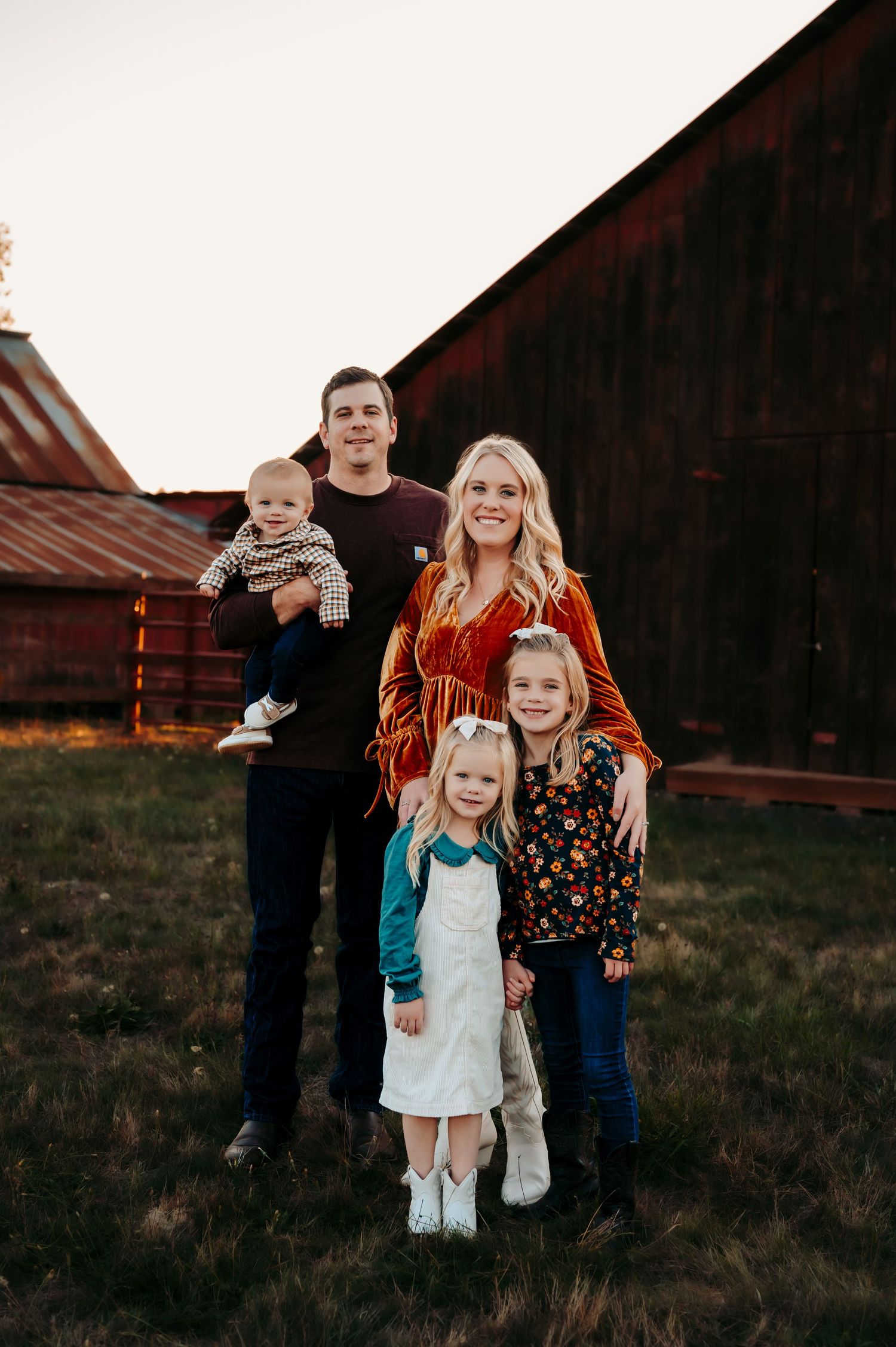 Family of five poses together for portrait in front of weathered red barn at sunset.