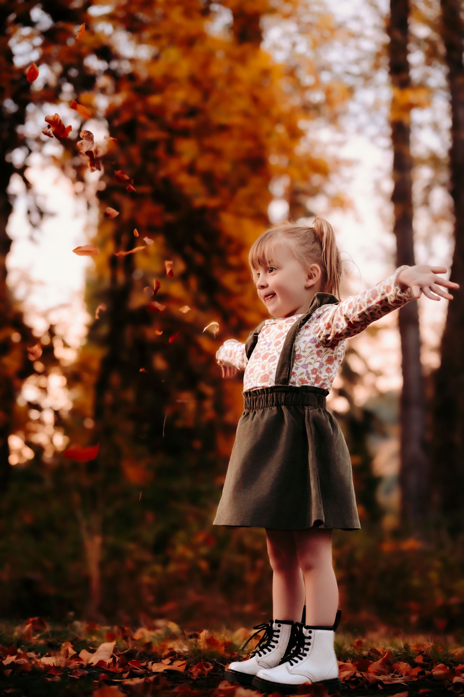 Young child in a dress and boots plays joyfully with falling autumn leaves in a forest.
