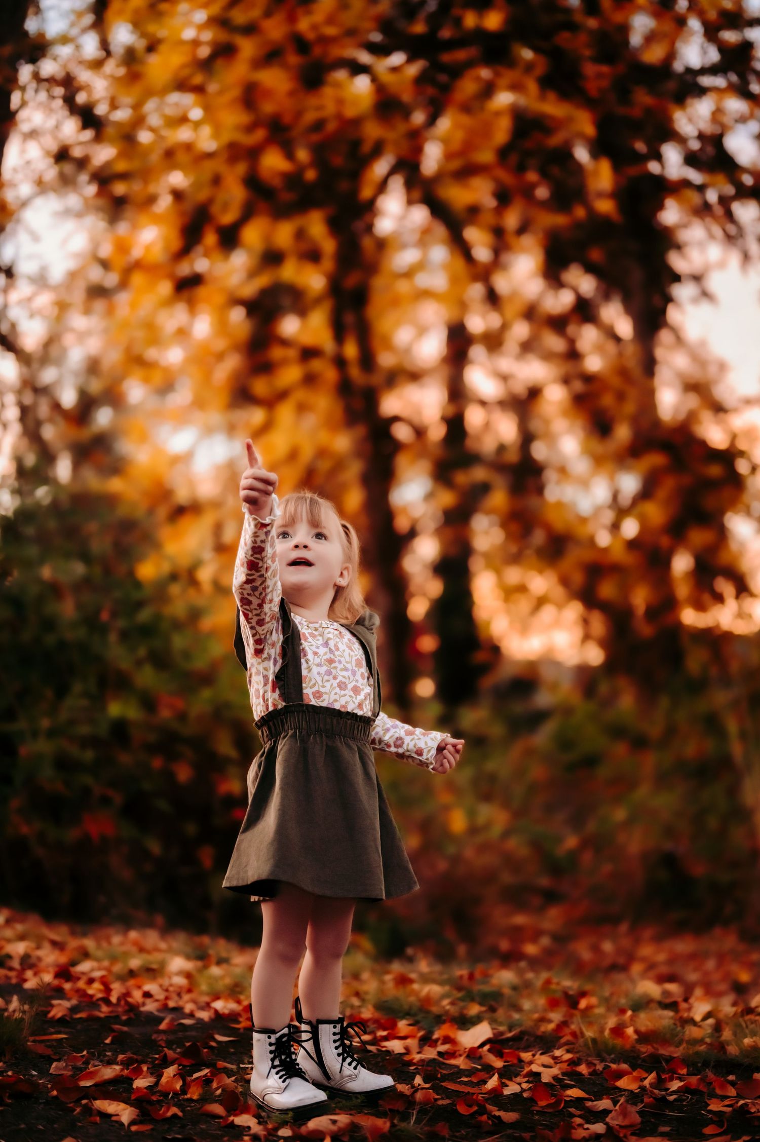 Child in a dress and boots reaches up towards falling leaves in an autumn forest scene.