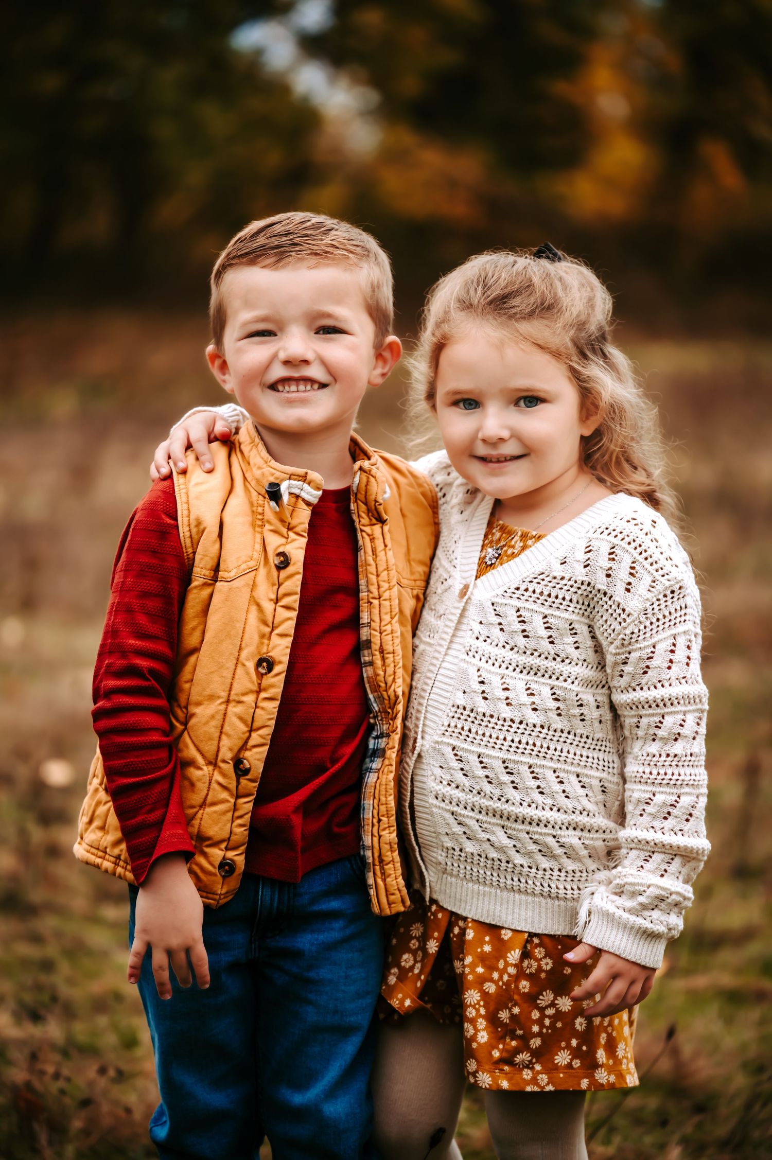 Two children in autumn outfits smile together outdoors.