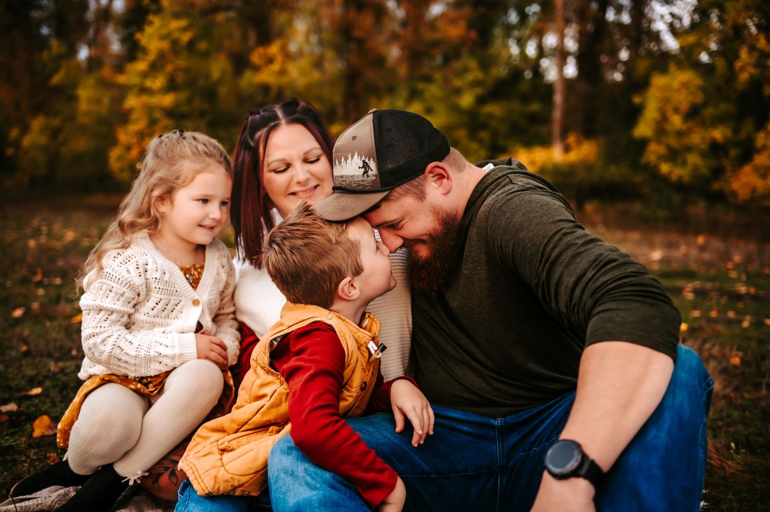 A family shares a tender moment together on fallen autumn leaves in a park setting.