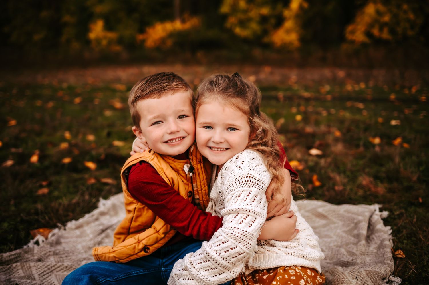 Two young children in sweaters hugging and smiling while sitting on a blanket surrounded by fall leaves.