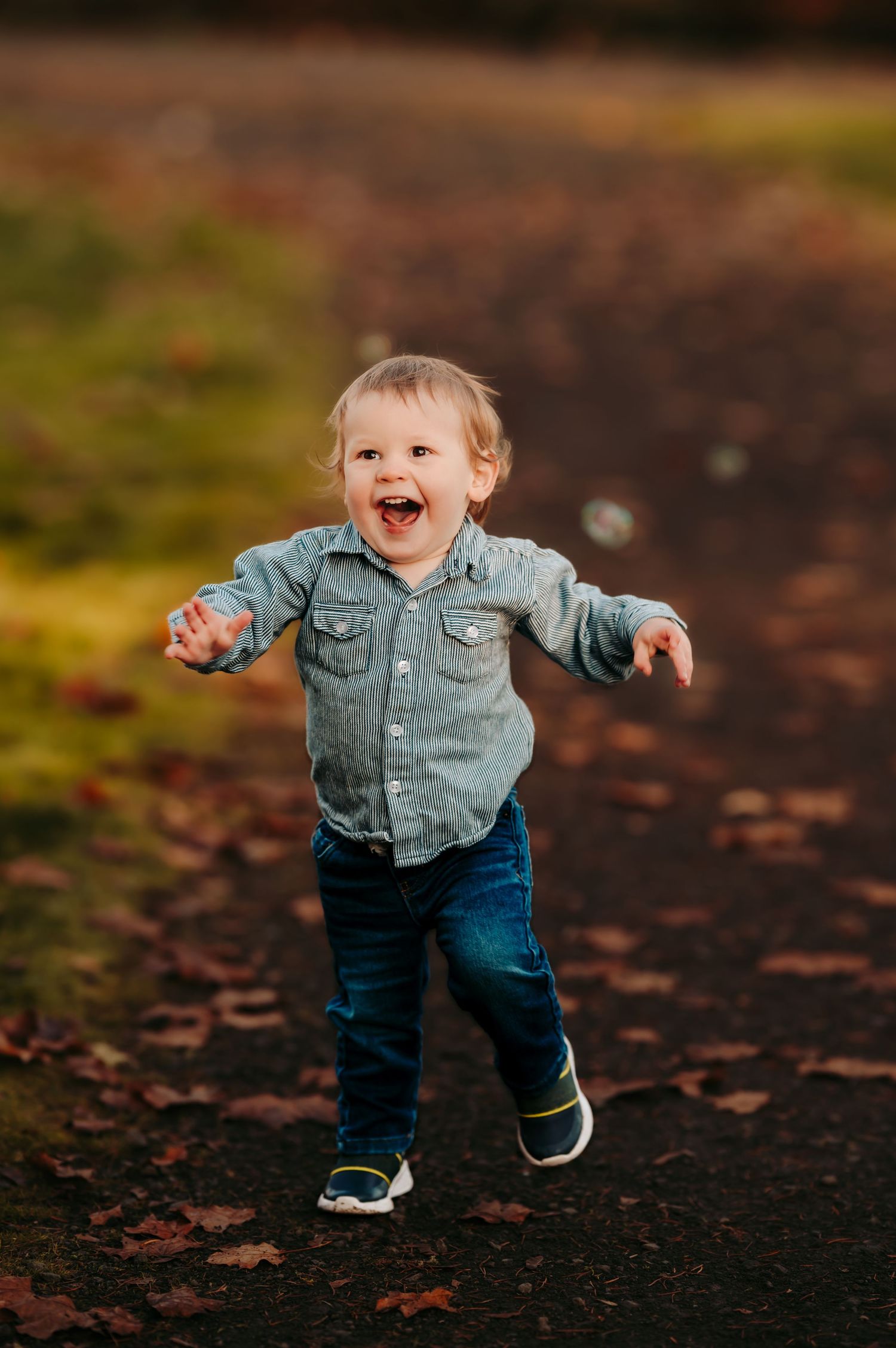 Toddler in jean jacket runs through fallen leaves on autumn path.