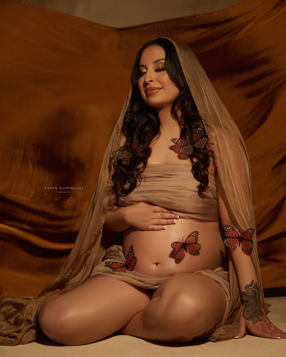 Maternity photo in warm tones featuring a pregnant woman in flowing veil with decorative butterflies against brown fabric backdrop.