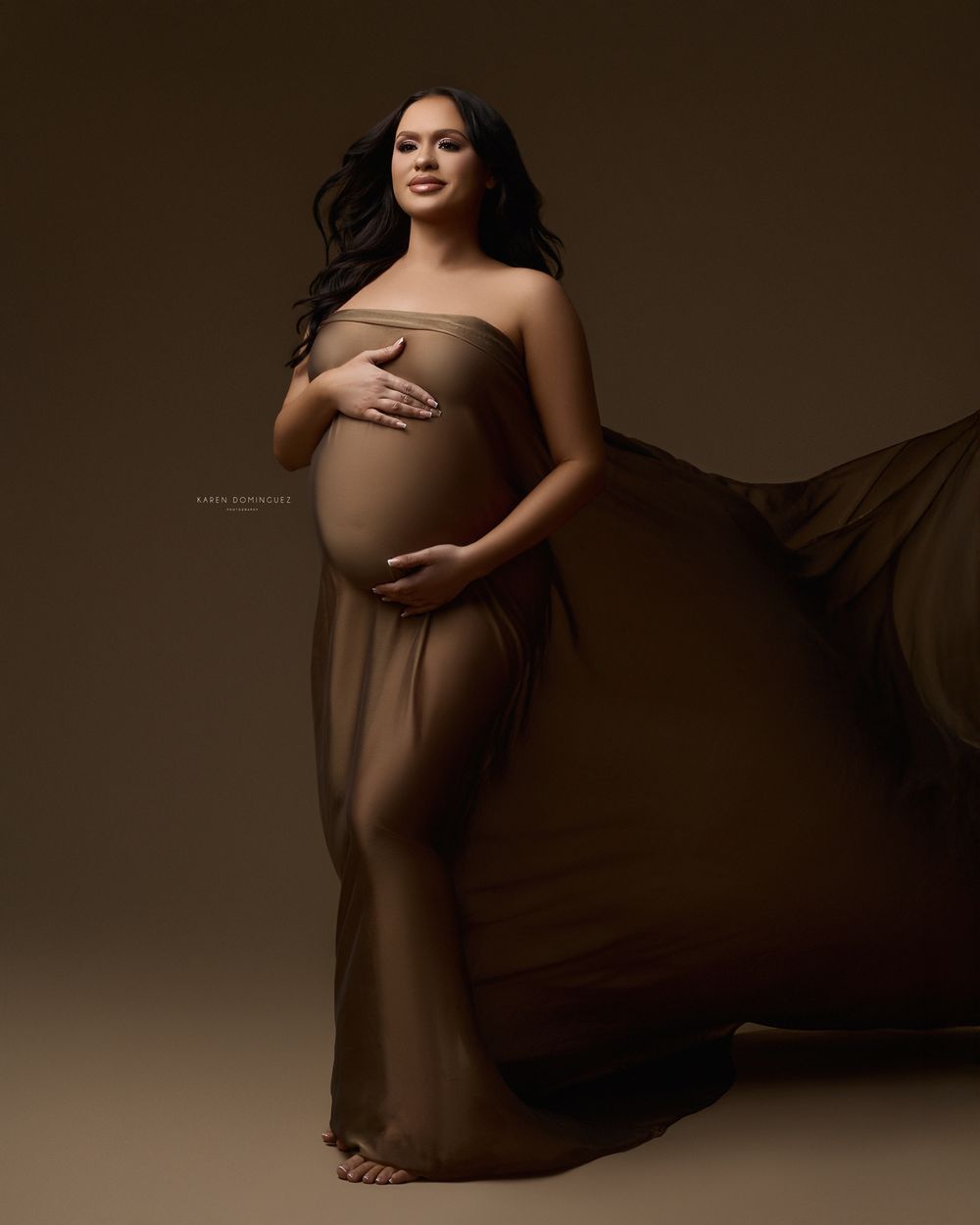 Professional maternity photoshoot in warm brown tones with flowing fabric and artistic lighting.