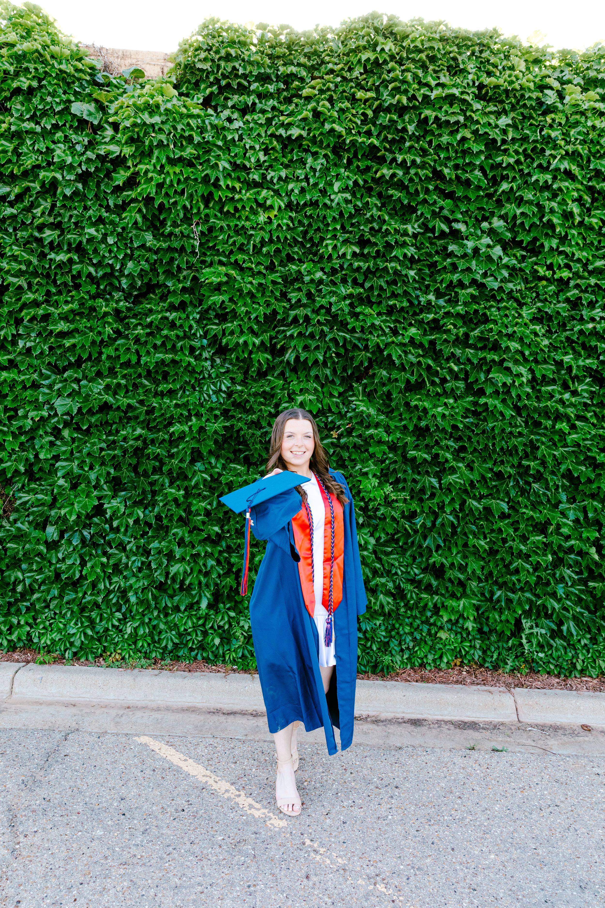 Graduate in blue academic regalia poses in front of lush green ivy wall.