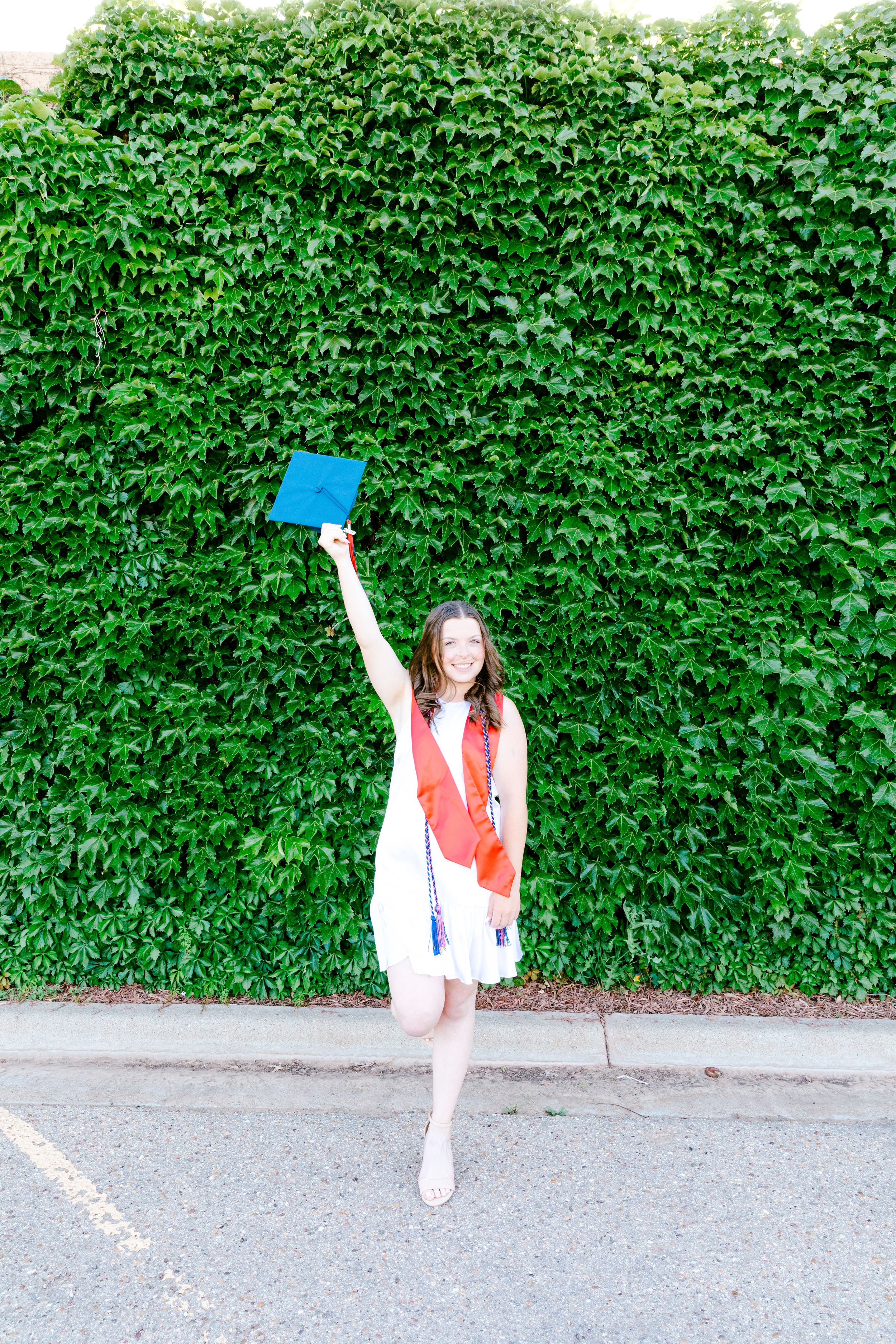Graduate in white dress and red stole holds up blue diploma against lush green ivy wall.