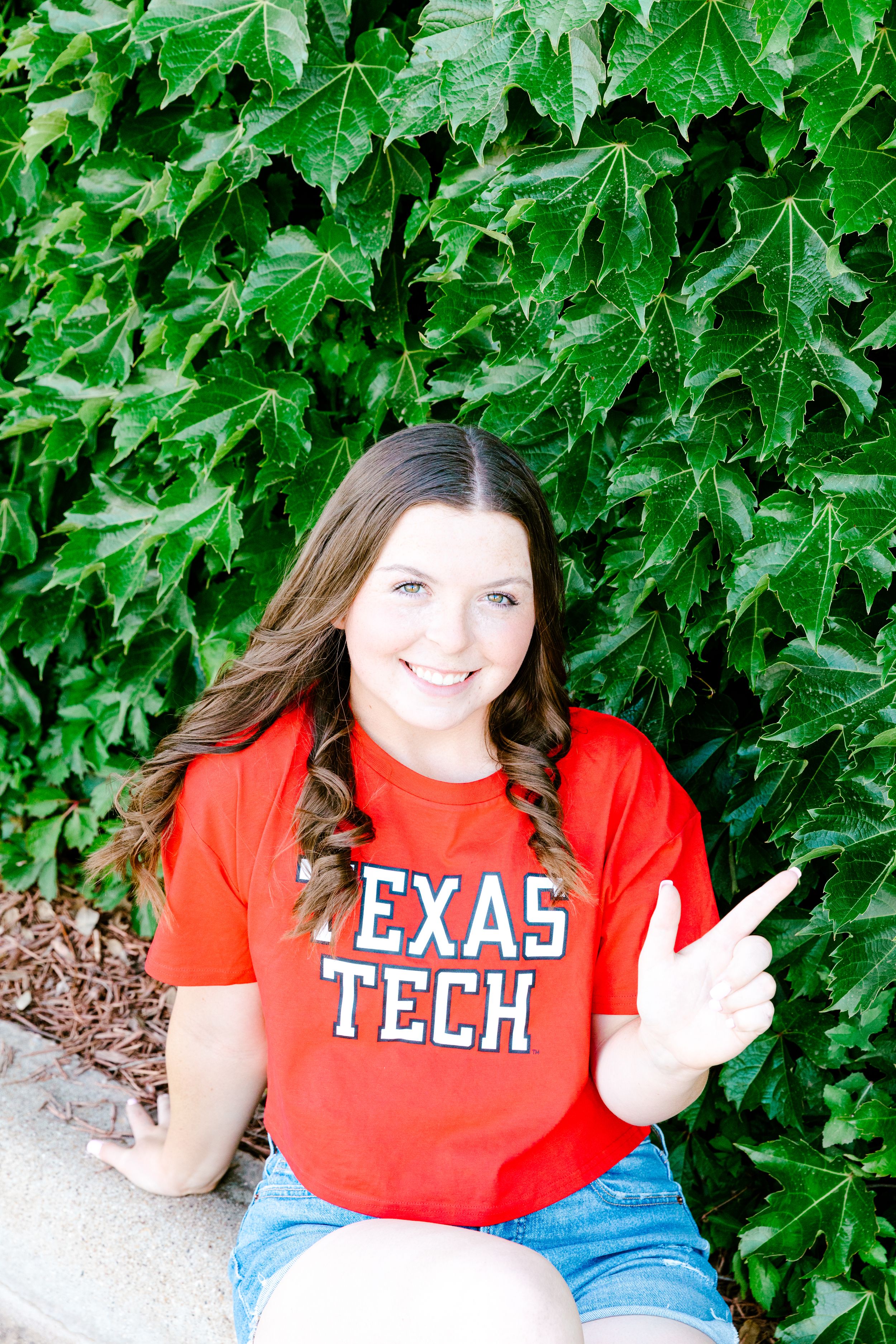 Student in red Texas Tech shirt and denim shorts poses against green ivy backdrop.