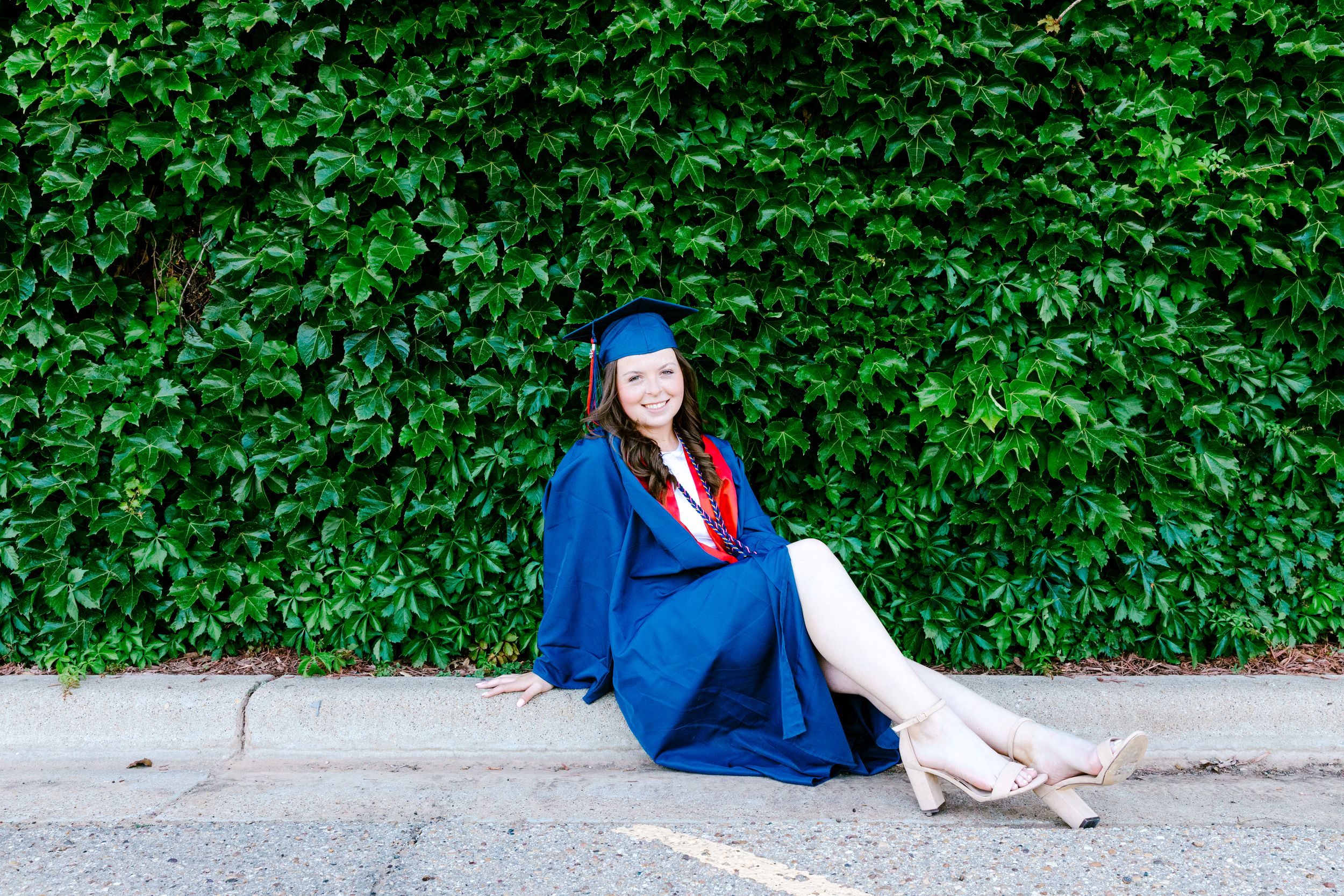 Graduate in blue cap and gown sits against ivy covered wall outdoors.