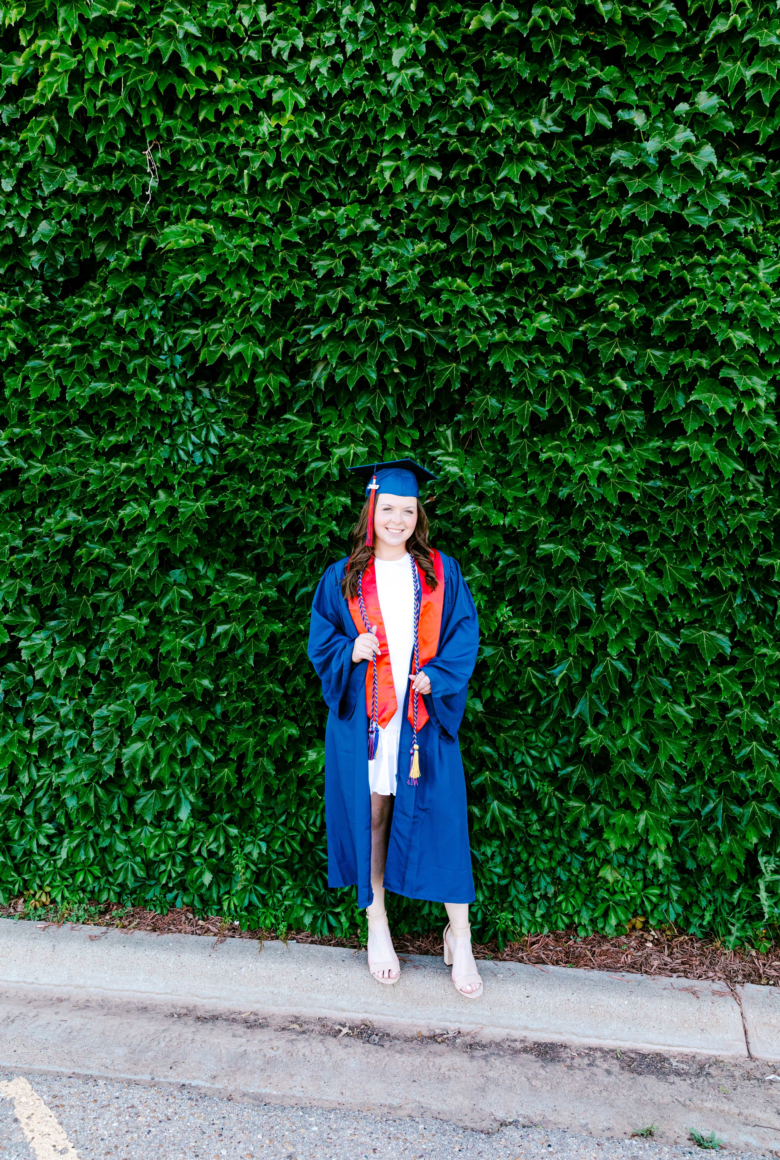 Graduate in blue cap and gown stands against wall of green ivy leaves.