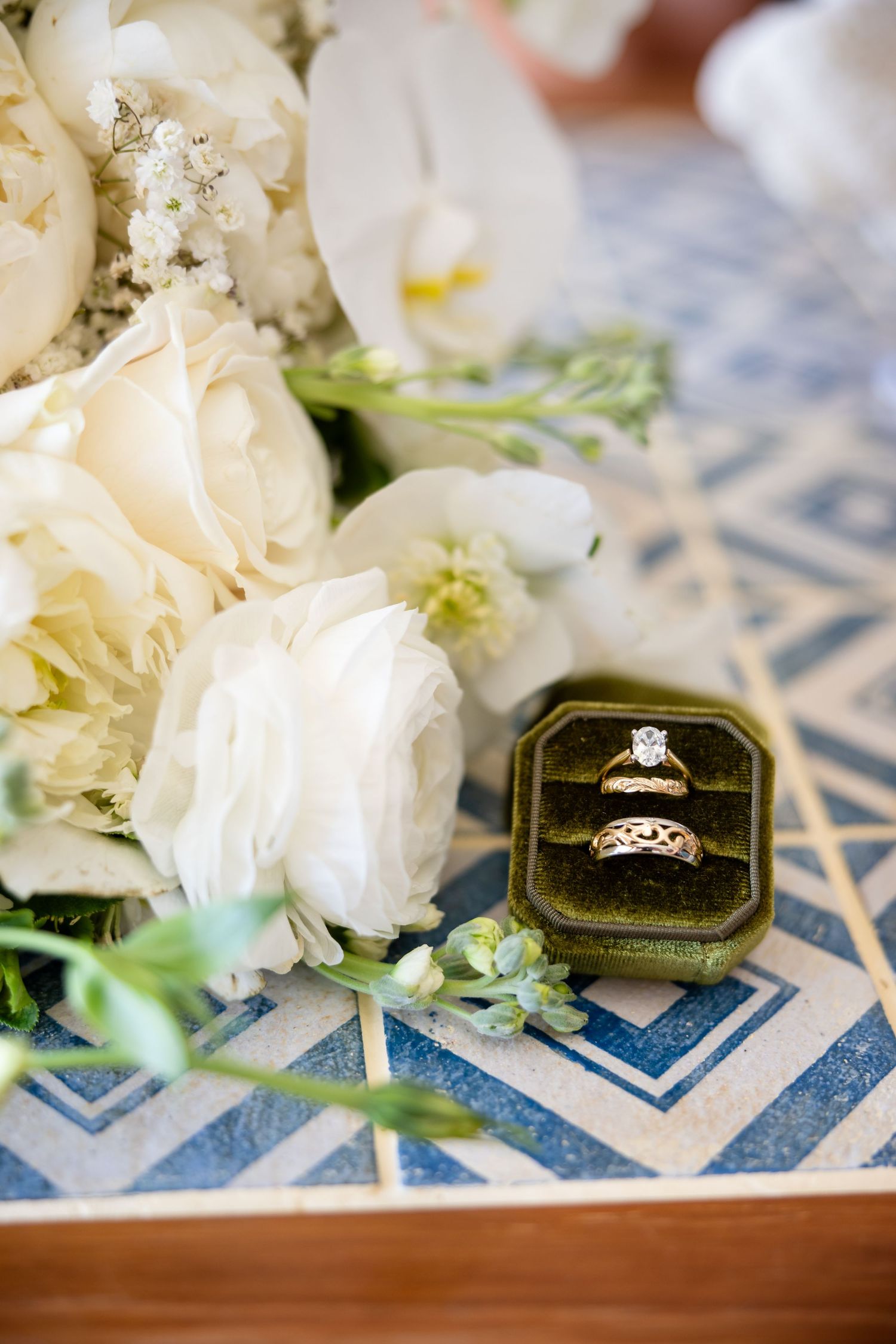 Gold wedding rings in a green velvet box displayed beside white roses and orchids on a blue geometric surface.