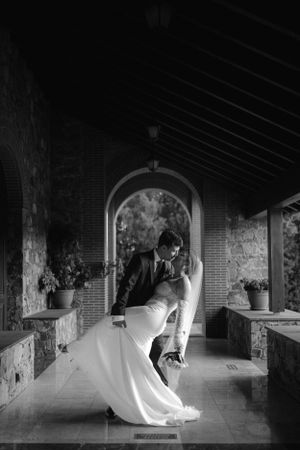 A romantic black and white photo of a couple dancing in an arched hallway with stone walls and potted plants.