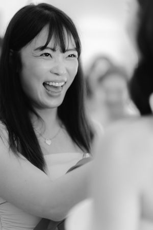 A black and white portrait of someone smiling joyfully with long dark hair and bangs wearing a light colored top.