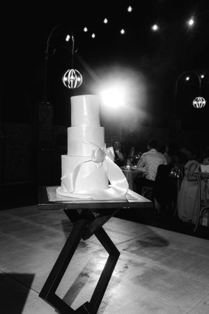 An elegant white wedding cake displayed on a modern geometric stand under glowing pendant lights at an evening reception.