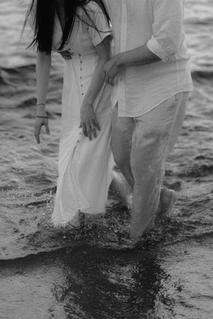 A romantic black and white photo of a couple standing in shallow water wearing white clothing at the beach.