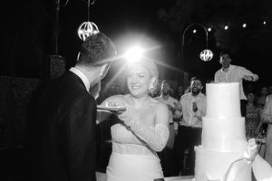 A couple shares their first dance at an evening wedding reception with a white wedding cake visible in the background.