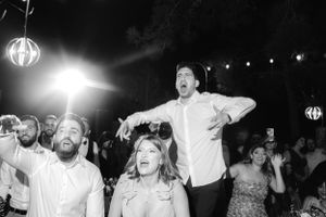 Black and white photo of wedding guests dancing energetically under a glowing pendant light at a reception celebration.