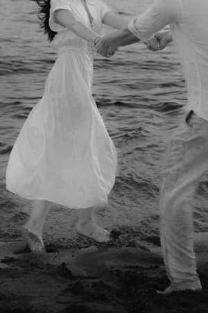 A couple dances together on the beach, with the woman's white dress flowing in the breeze against dark water.