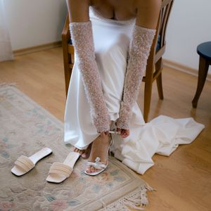 A pair of elegant lace bridal gloves and white sandals arranged on a patterned rug with wedding dress details visible.