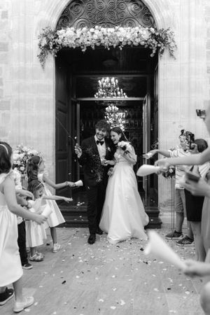 A wedding couple exits through an ornate doorway decorated with flowers as guests throw petals in black and white photography.