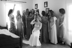 A group of women in formal dresses gather in a sunlit room as a person in a white gown sits in the center.