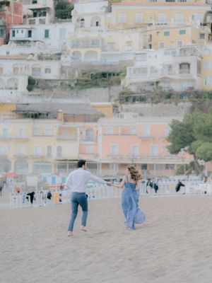 Two people running playfully on a sandy beach with colorful pastel buildings of Positano, Italy cascading down the hillside.