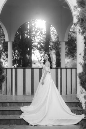 Black and white photograph of an elegant long wedding dress with a flowing train under an arched gazebo.