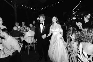 A black and white photo of a couple walking through a reception venue with string lights overhead and guests seated nearby.