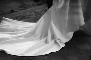 A flowing white wedding dress train drapes elegantly across a dark wooden floor in black and white photography.