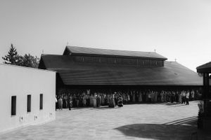A black and white photograph shows a crowd of people gathered outside a large wooden building with a sloped roof.
