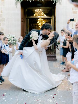 A romantic dip kiss between newlyweds outside a church entrance as guests shower them with flower petals.